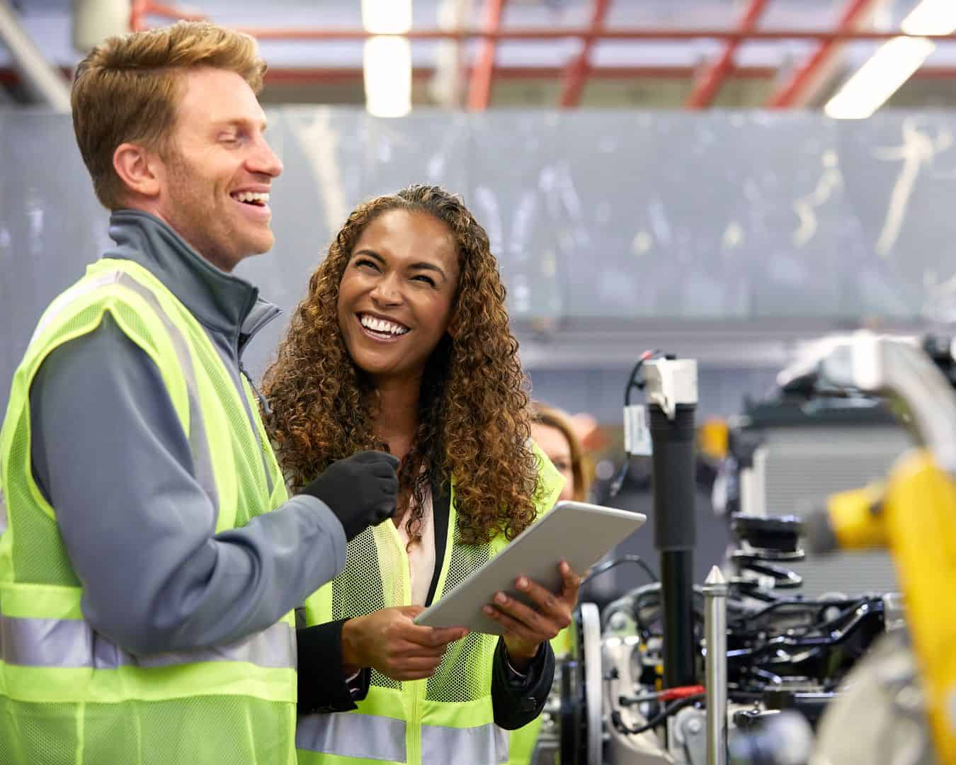 A joyful moment captured between two team members in high-visibility vests on the manufacturing floor, as they review data on a tablet, reflecting the positive team dynamics and quality-centric culture vital for industrial excellence.